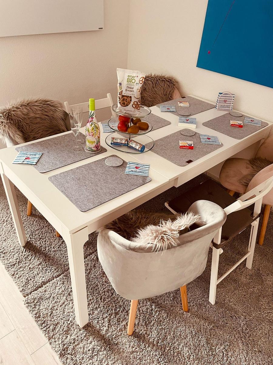 Weißer Tisch mit Stühlen, Platzdeckchen und Obst. Wandbild und Teppich sichtbar.