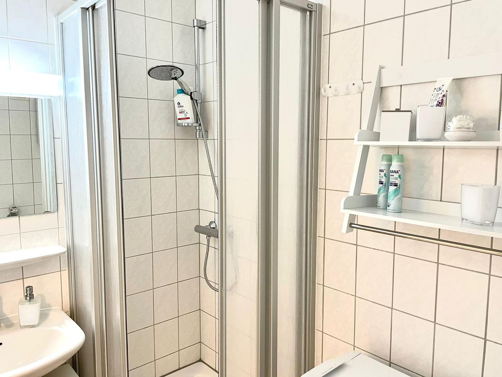 Badezimmer mit Dusche, Waschbecken und Regal. Fliesenwand und Spiegel sichtbar.