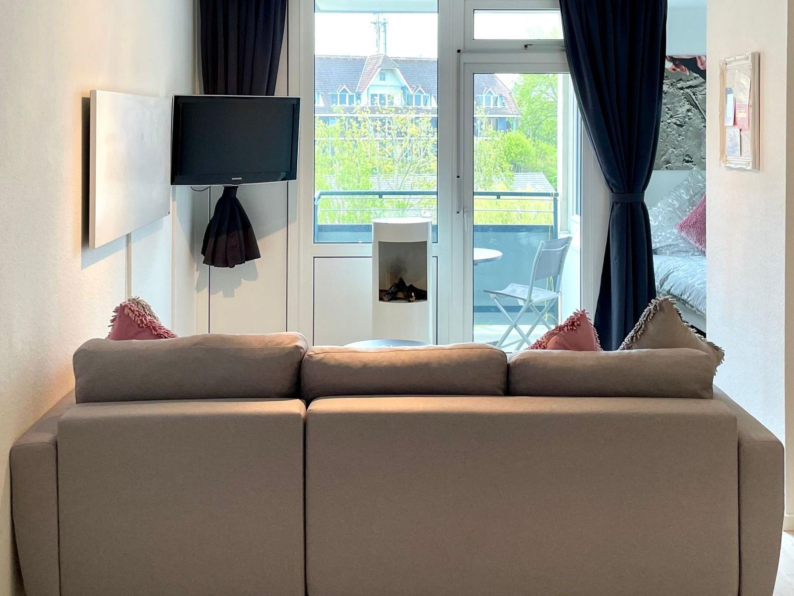 Ein Sofa mit Kissen vor Fenster und Balkon. Fernseher an Wand. Blick auf Haus und Grün.
