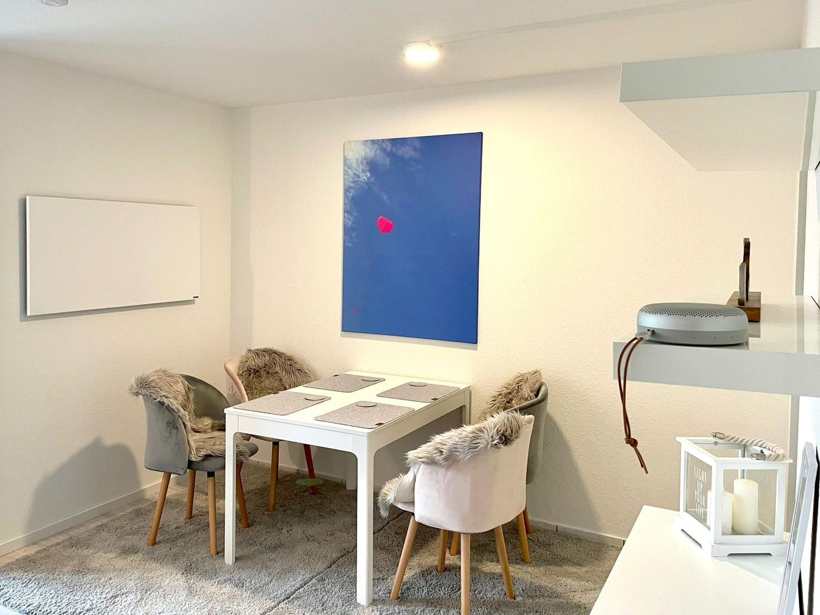 Essbereich mit weißem Tisch, vier Stühlen und Wandbild mit Herz. Hintergrund: weiße Wände und Decke.