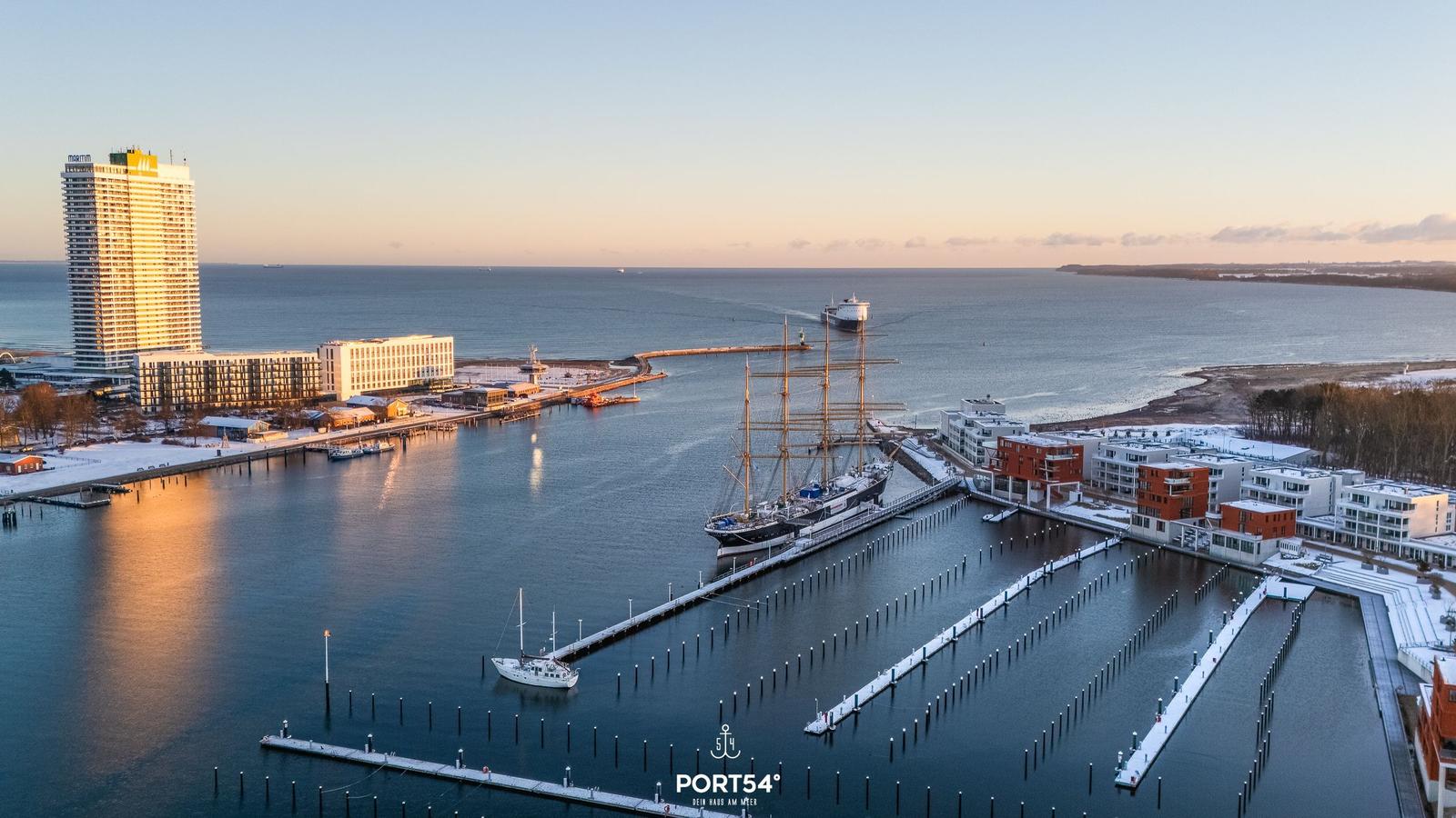 Hafen mit Segelschiff, modernen Gebäuden und ruhigem Wasser bei Sonnenuntergang.