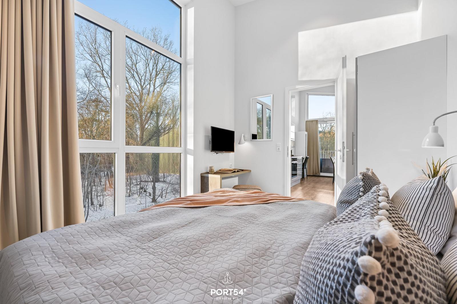 Schlafzimmer mit großem Fenster, Bett und Blick auf Wald. Küche und Bad sind angebunden.