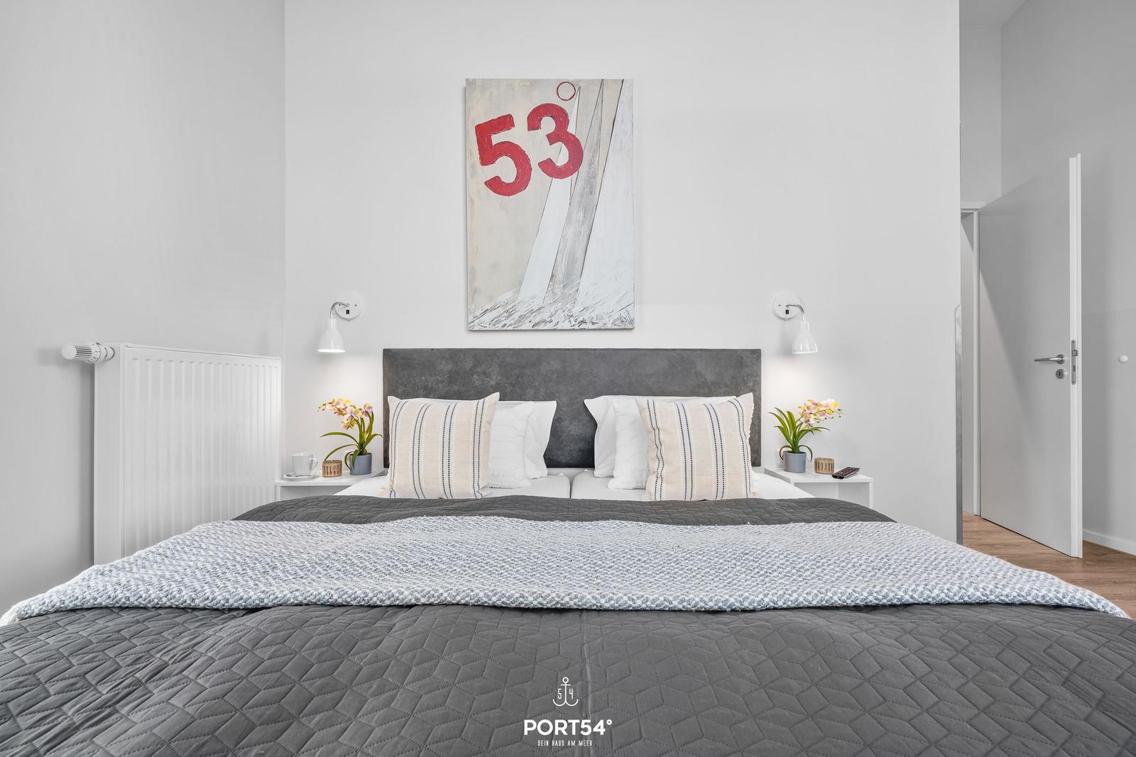 Schlafzimmer mit Bett, Kopfkissen und Wandbild mit der Zahl 53.