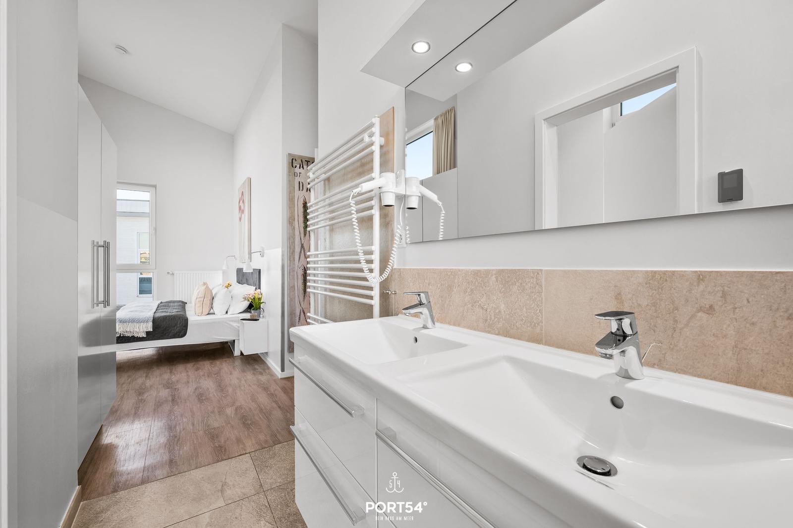 Badezimmer mit Doppelwaschbecken und Spiegel. Im Hintergrund ist ein Schlafzimmer mit Bett und Fenster.