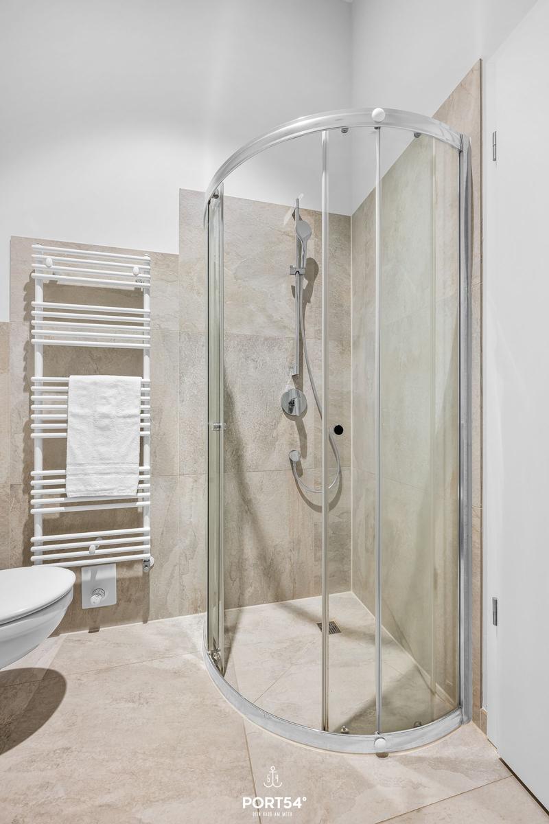 Duschkabine mit Glaswand, Handbrause und Heizung. Toilette und Handtuchhalter sichtbar.