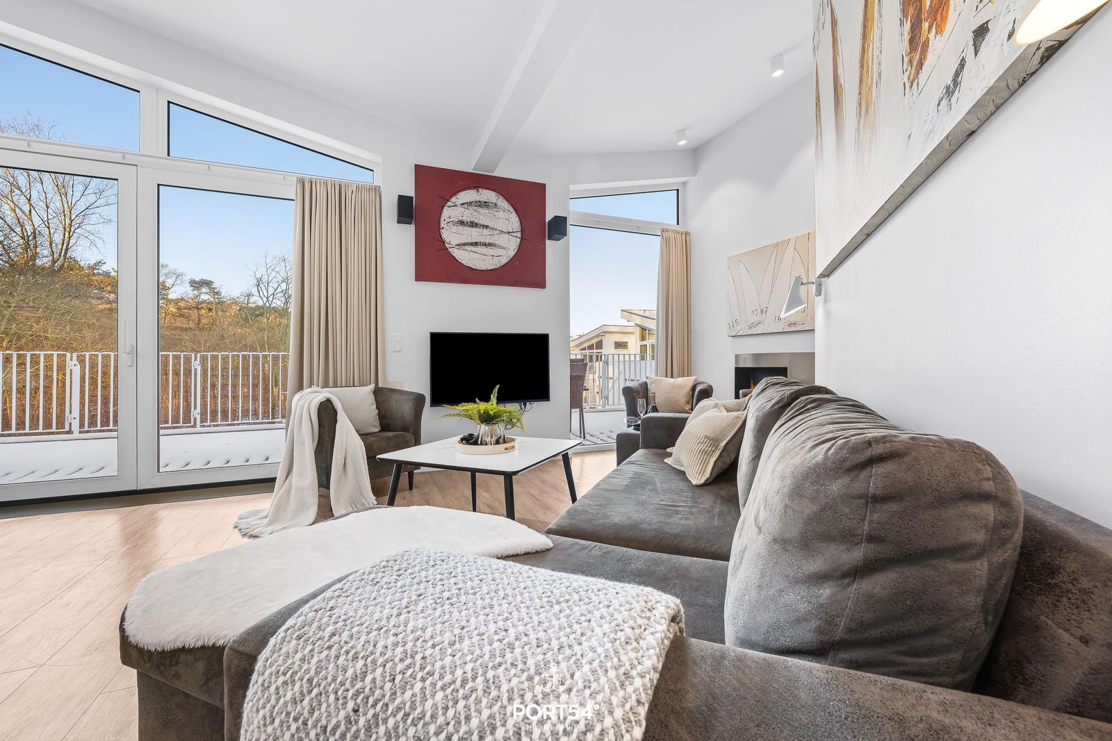 Wohnzimmer mit Sofa, Couchtisch und Fernseher. Große Fenster und Balkon mit Blick auf Wald.