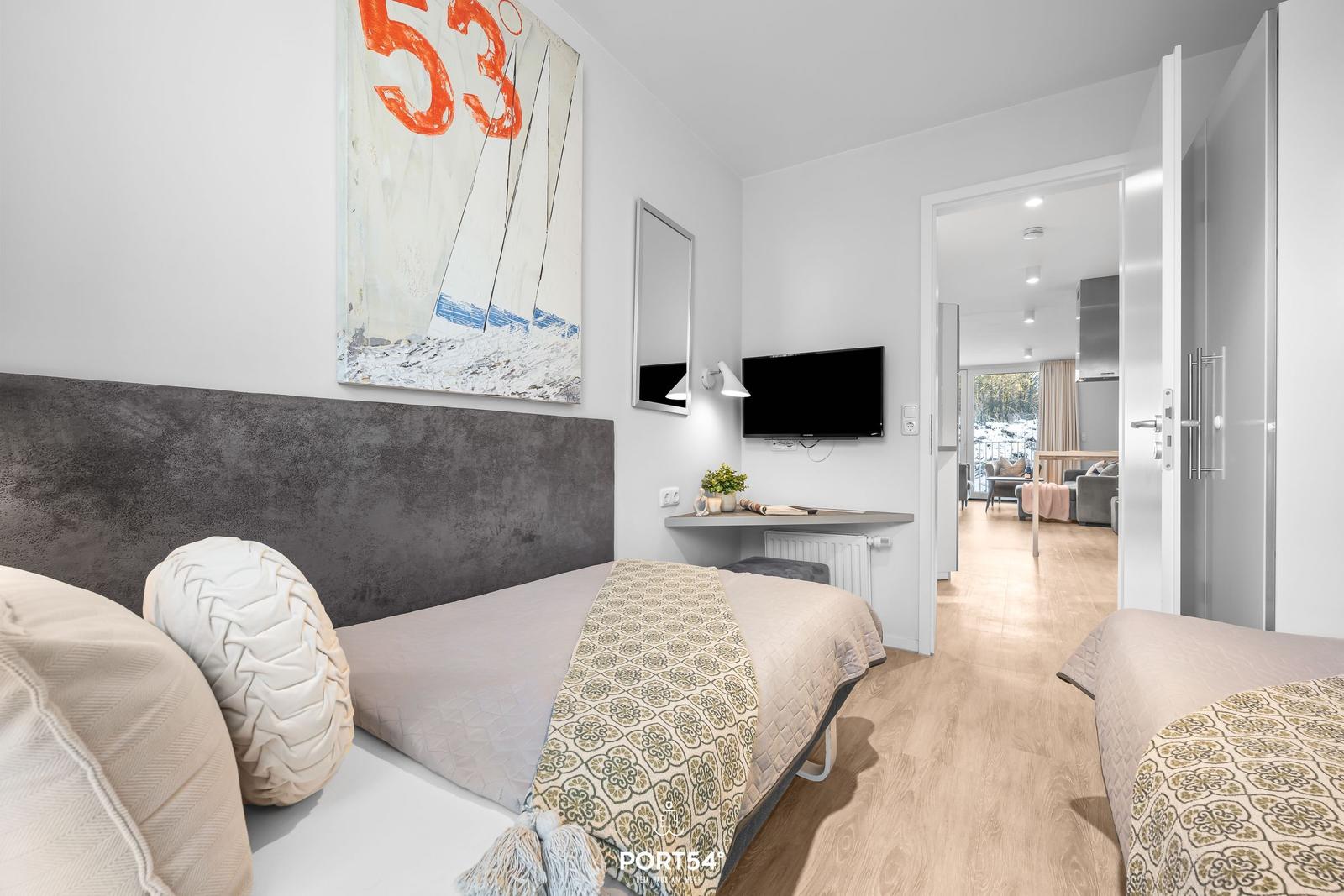 Zwei Betten mit Musterdecken, Wandbild und TV. Durchgang zur Wohnküche mit Balkon.