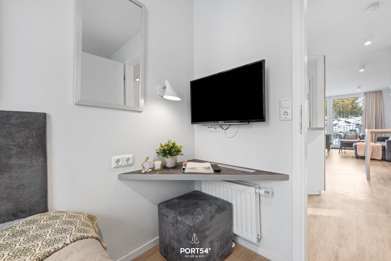 Schlafzimmer mit Fernseher, Spiegel und Schreibtisch. Blick in Wohnbereich mit Sofa.