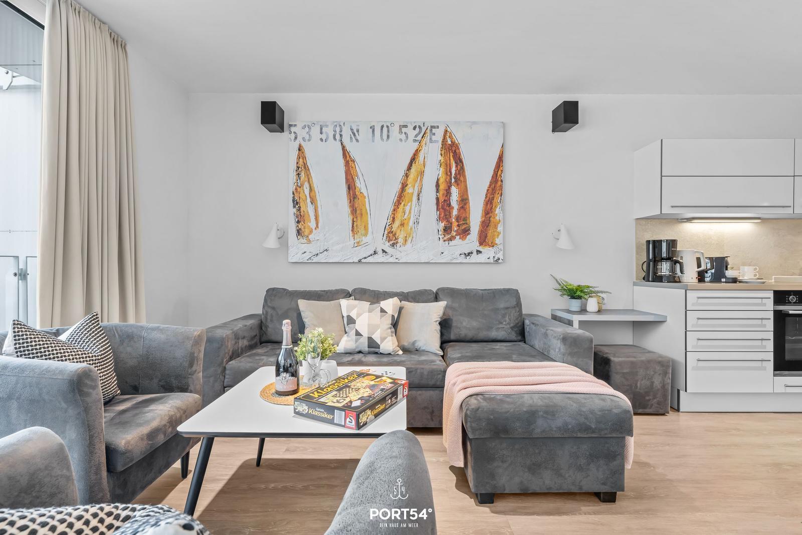 Wohnzimmer mit grauem Sofa, Küchenbereich und Wandbild mit Segelbooten.