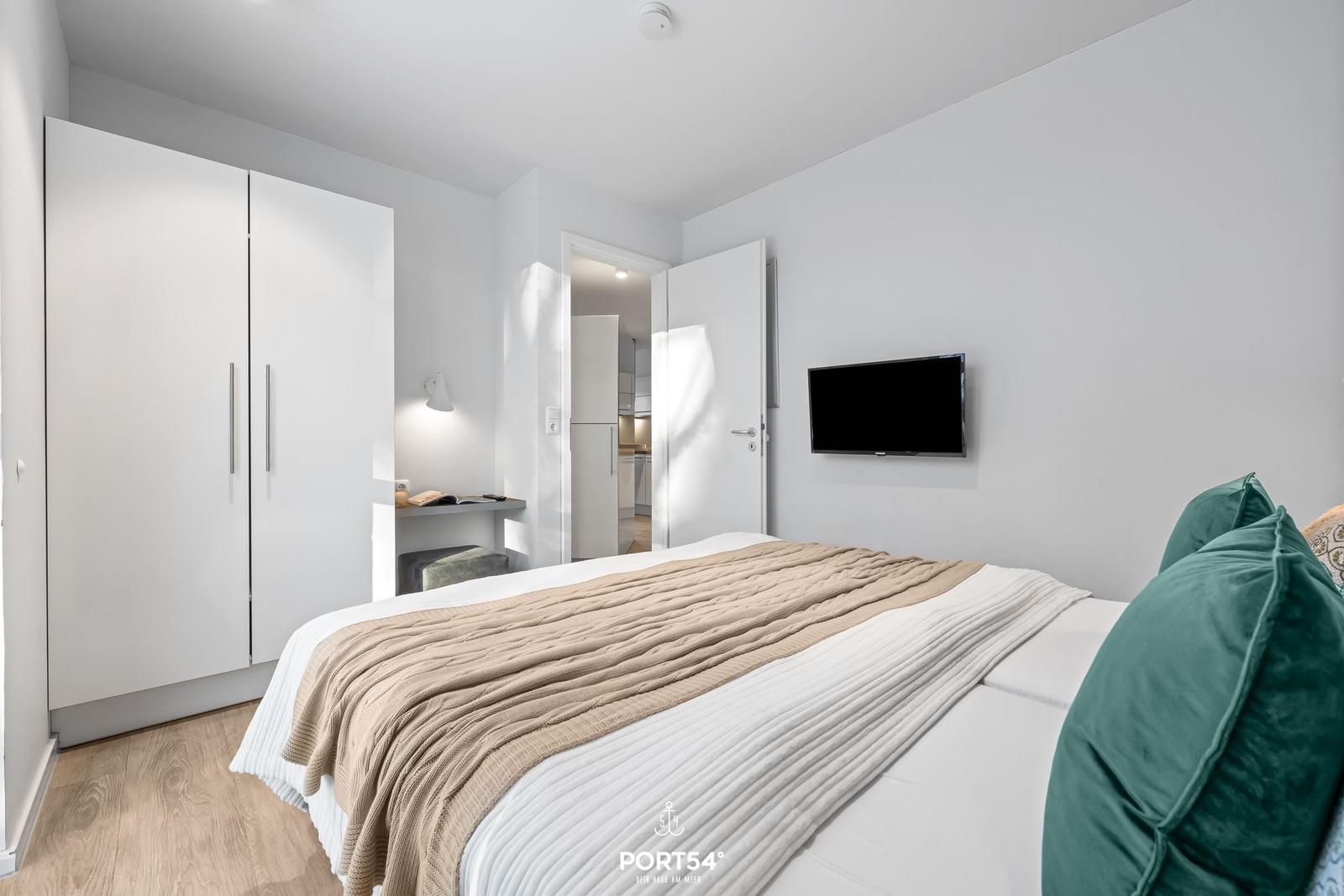 Schlafzimmer mit Bett, Kleiderschrank und Fernseher. Durchgang zur Küche sichtbar.