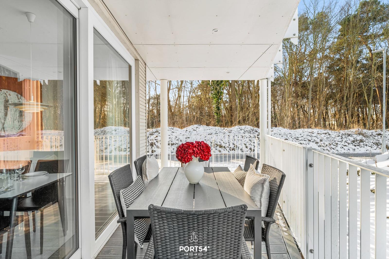 Terrasse mit Esstisch, Stühlen und roten Blumen. Schnee bedeckt den Boden und die Bäume.