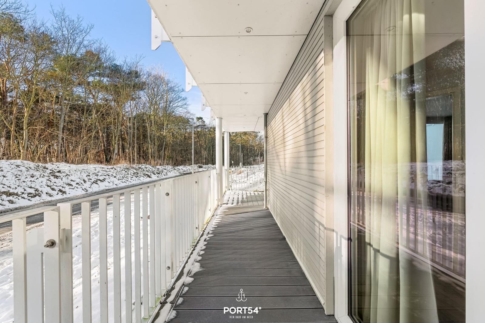 Längliche Veranda mit weißem Geländer und Holzdielen. Schnee bedeckt den Boden und die Bäume im Hintergrund.