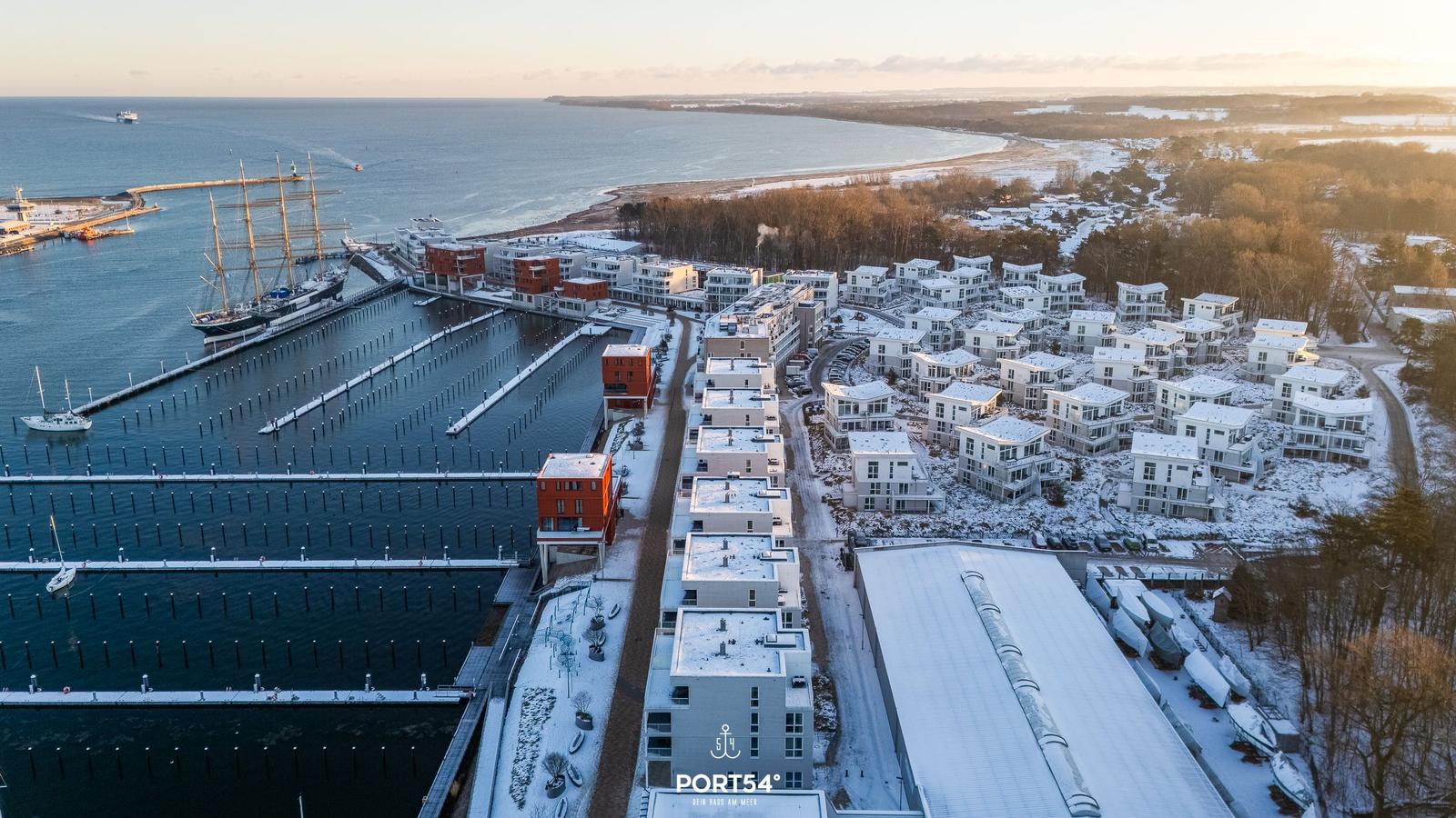 Schneebedeckte Häuser am Hafen mit Segelschiff und Blick aufs Meer.