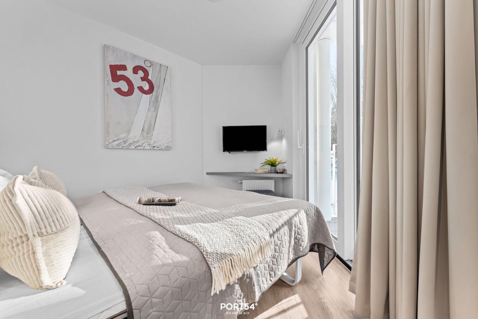 Schlafzimmer mit Bett, Wandbild und TV. Große Fenster mit Vorhängen.