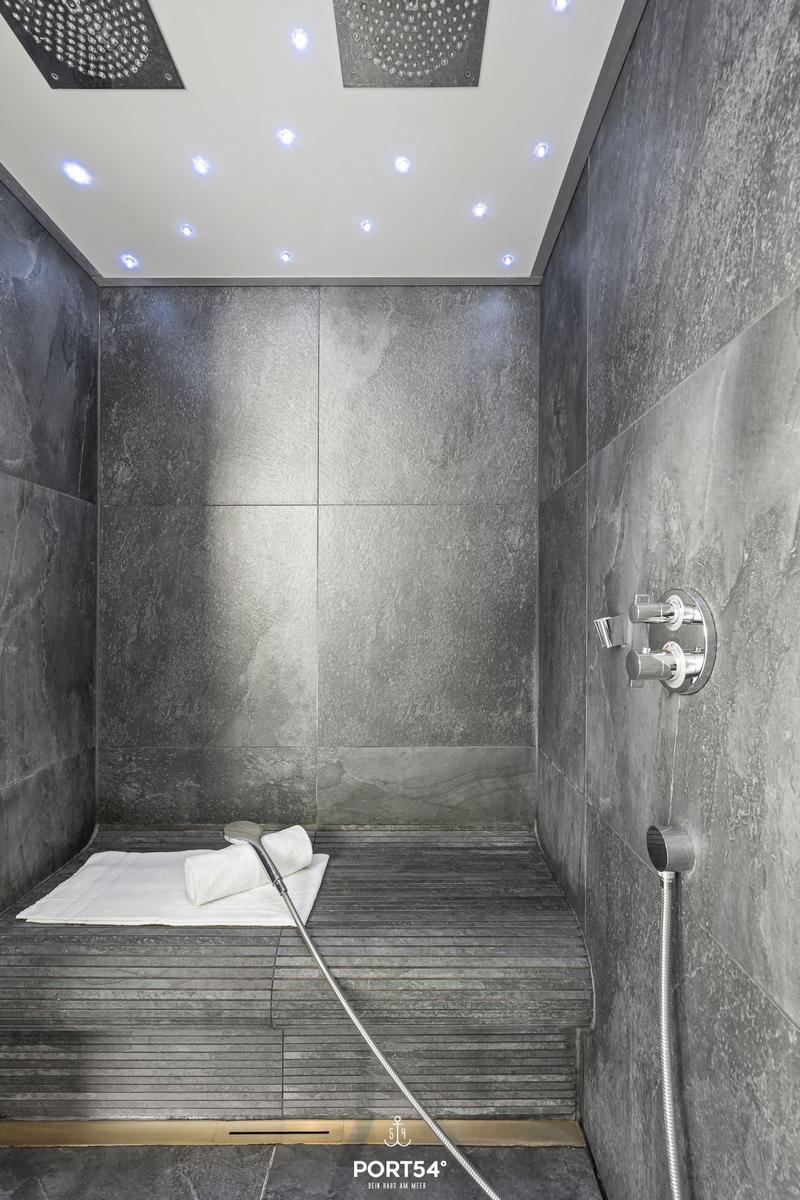 Duschkabine mit dunklen Fliesen, Duschkopf und beleuchteter Decke.