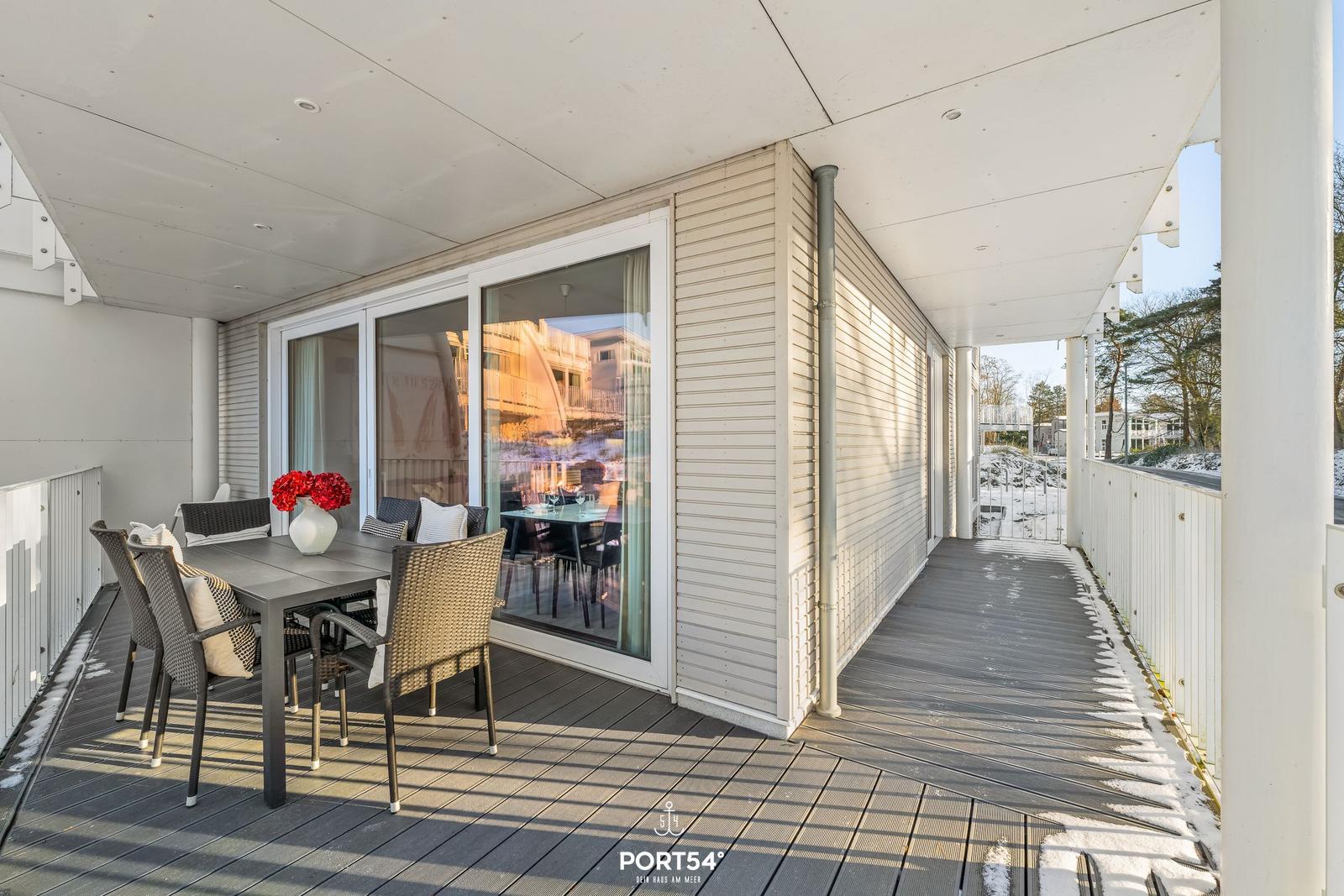 Terrasse mit Tisch, Stühlen und roten Blumen. Durchgehende Glasfront zum Innenraum.