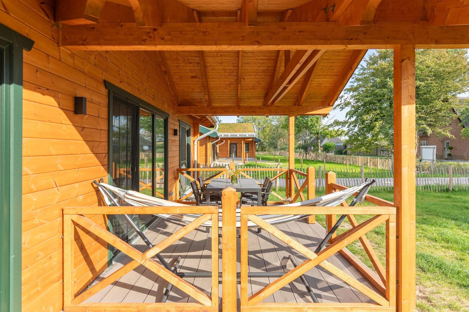 Terrasse mit Hängematten und Tisch unter Holzüberdachung. Blick auf grünes Gelände und Nebengebäude.