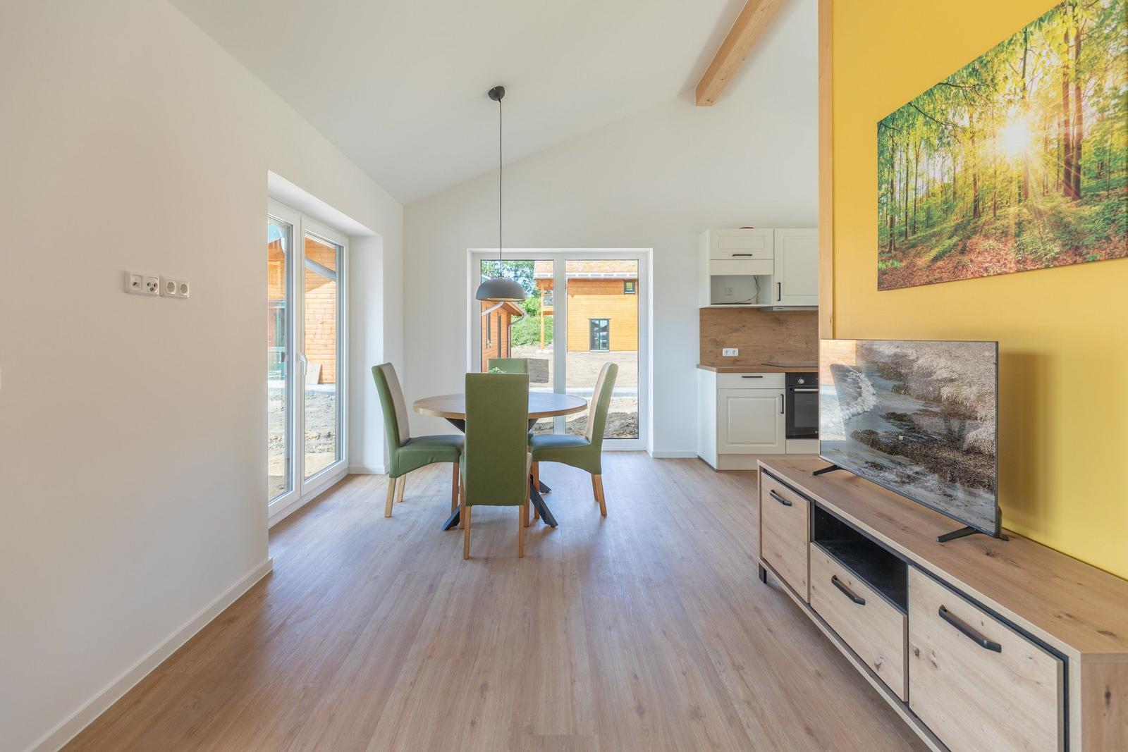 Wohn-Essbereich mit Esstisch, TV und Küche. Gelber Wandbereich mit Waldbild.