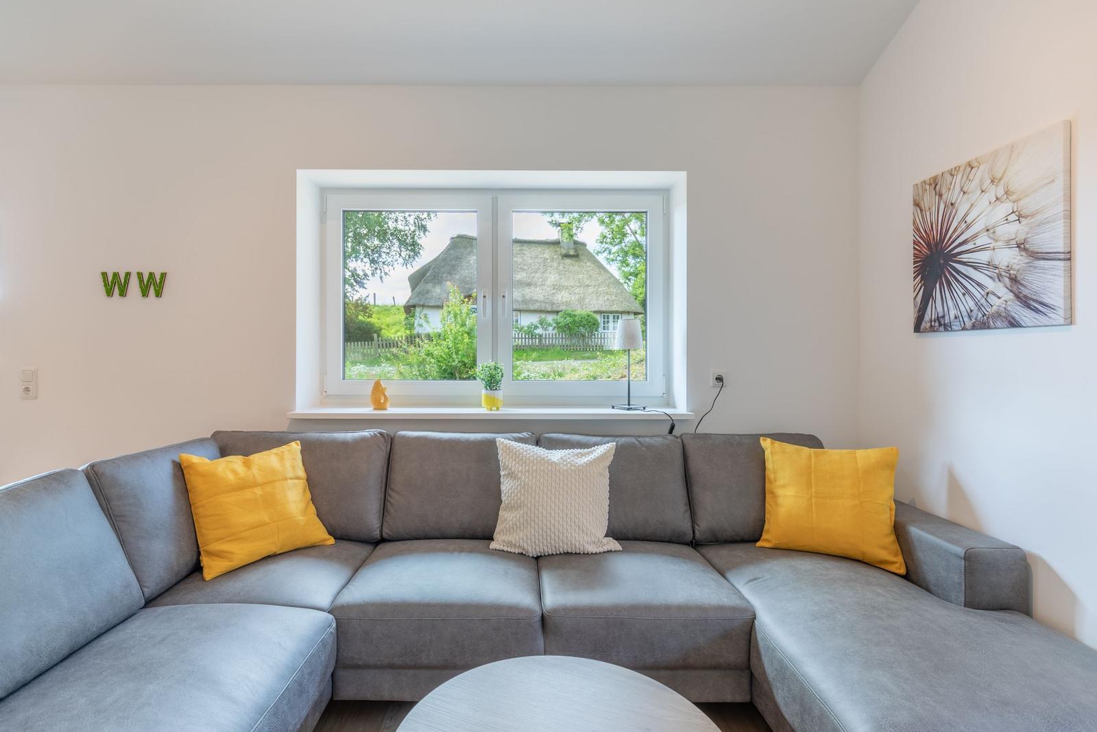 Ein grauer Ecksofa mit gelben und weißen Kissen vor einem Fenster mit Blick auf ein Strohdachhaus.