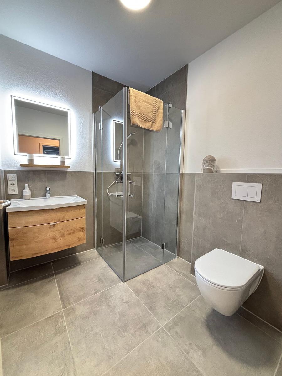 Bathroom with shower, sink, and toilet. Wooden vanity and lit mirror.