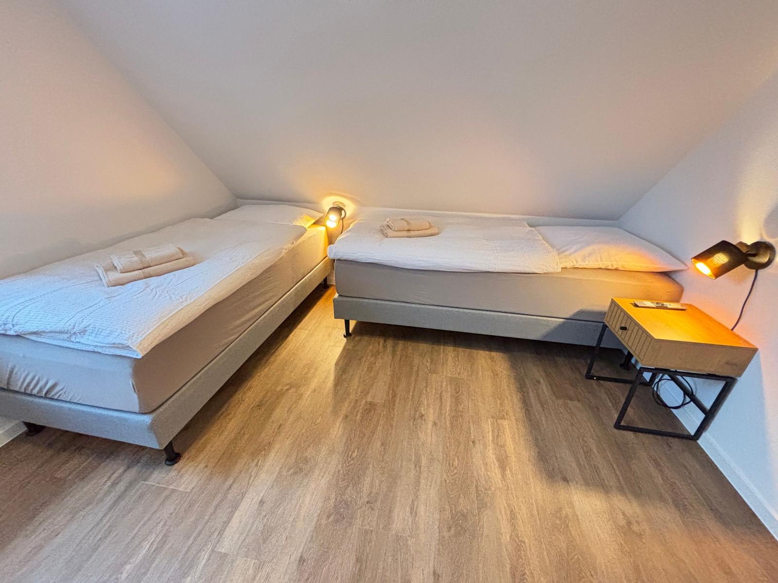 Two beds in an attic room with wooden floor and wall lamps.