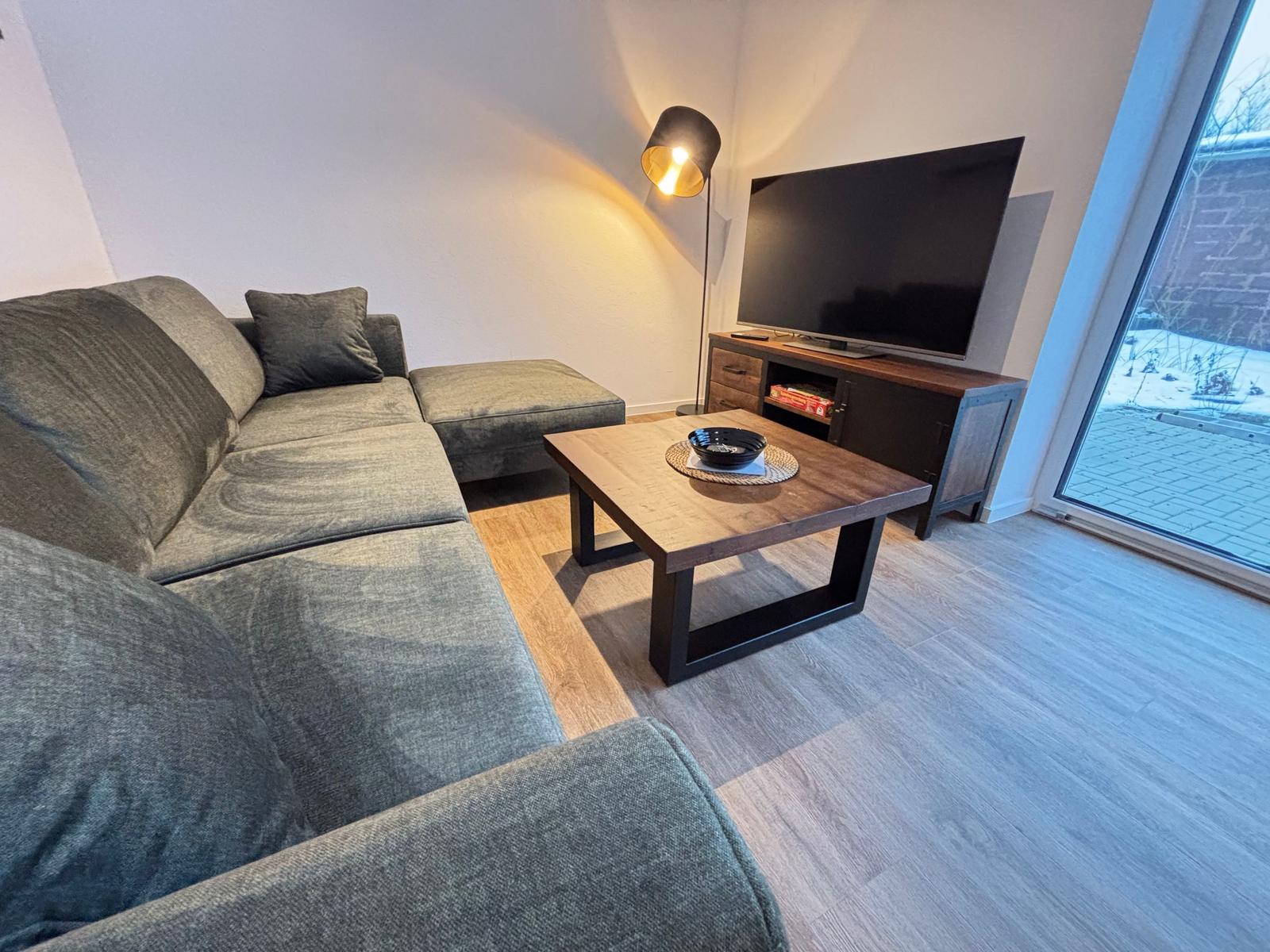 Living room with sofa, coffee table, and TV. Large window view to outdoor area.