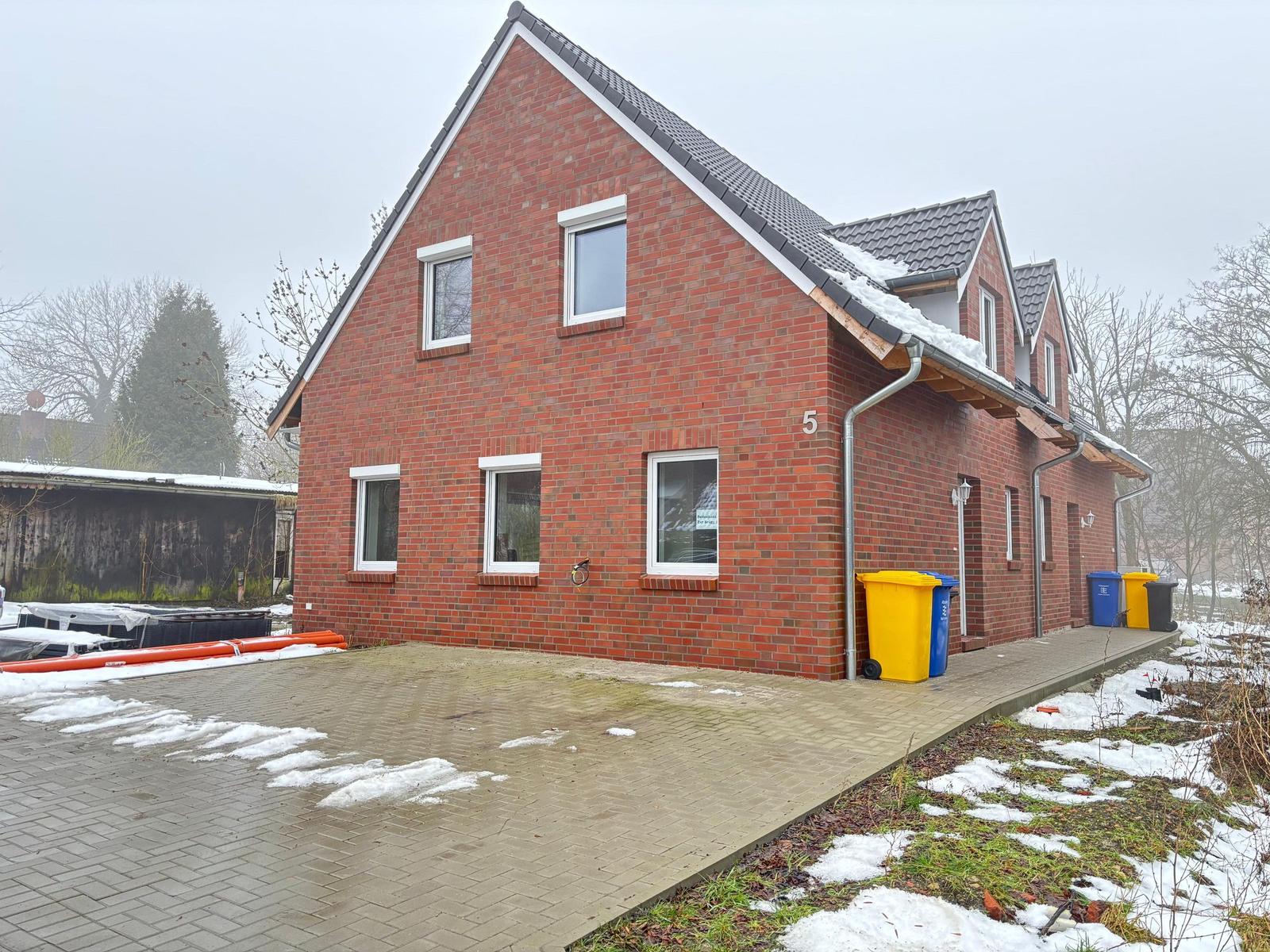 Red brick house with snow on roof and ground. Front side with windows and trash bins.