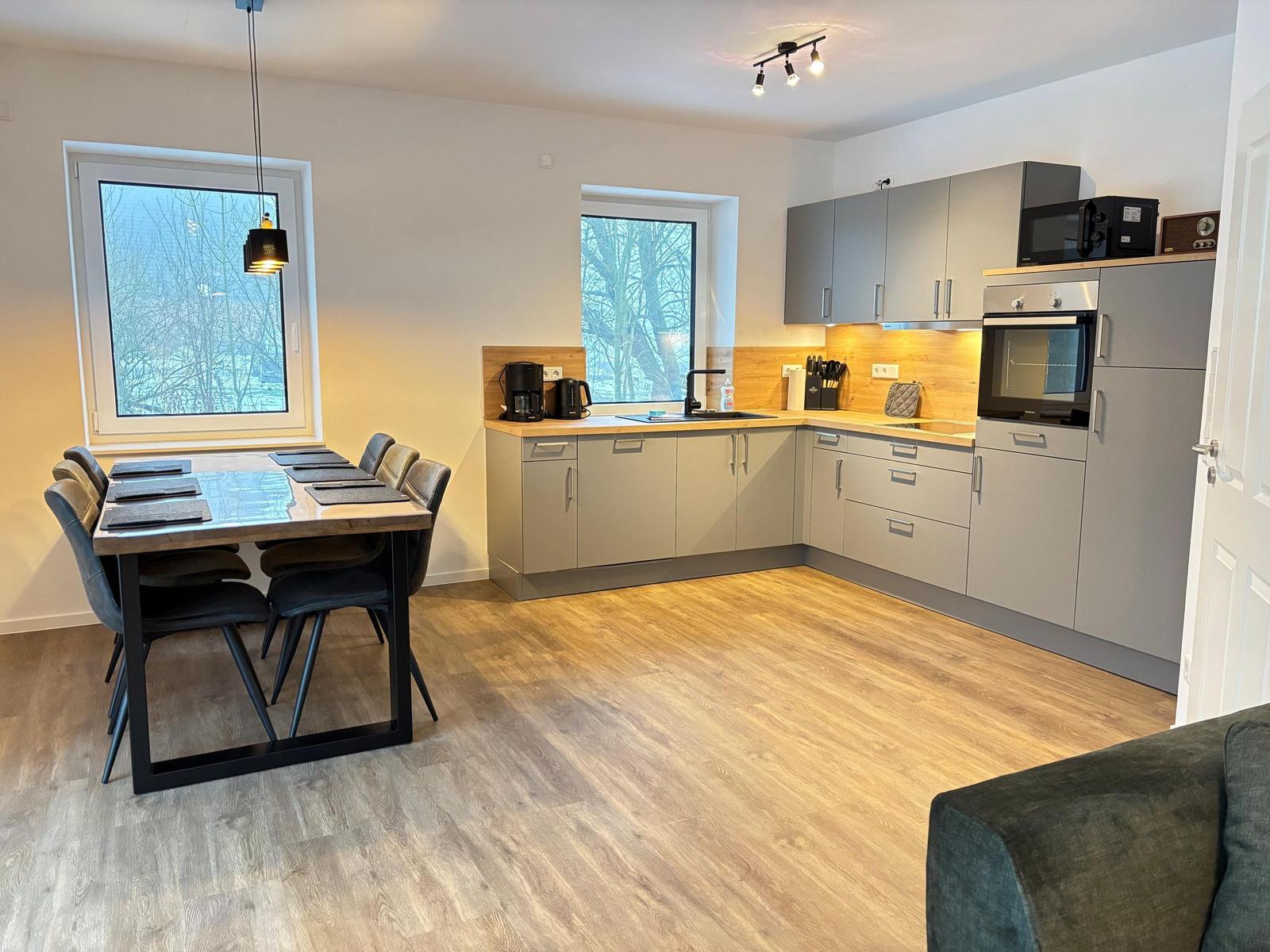 Kitchen with dining table, two windows, and wooden floor. Appliances and furniture are present.