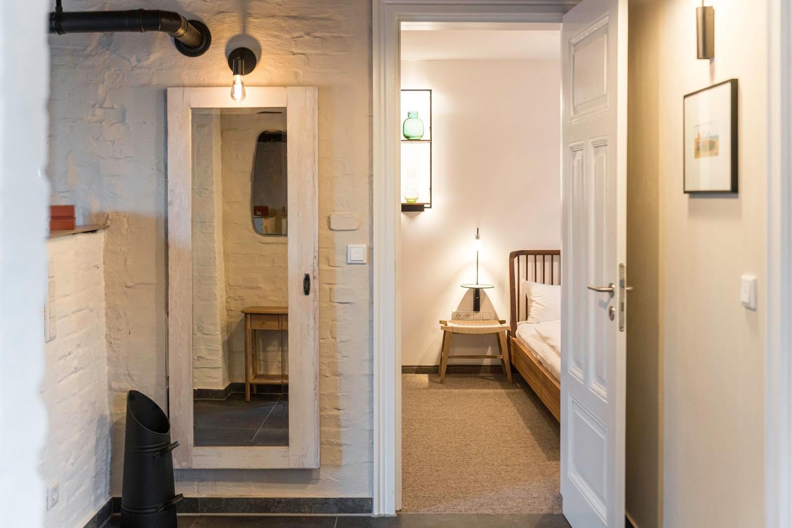 Flur mit Schlafzimmer und Spiegeltür. Bett, Nachttisch und Wandbeleuchtung sichtbar.