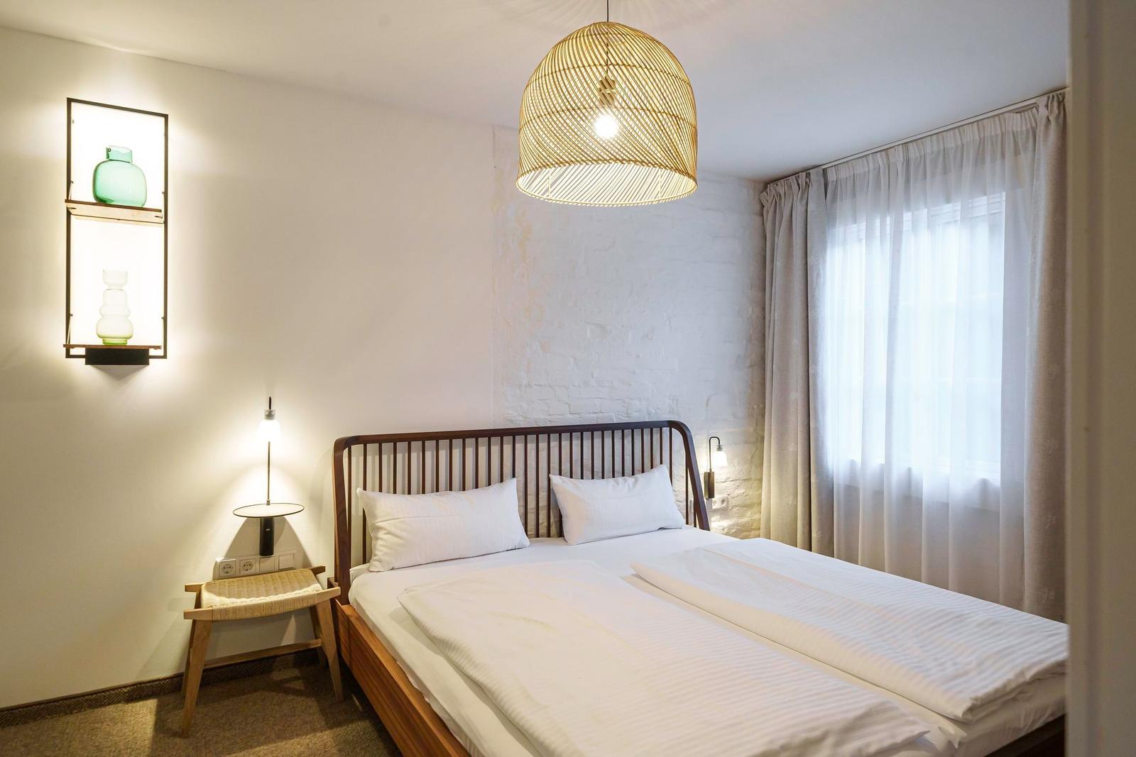 Schlafzimmer mit Doppelbett, Wandlampe und Deckenleuchte. Fenster mit Vorhängen.