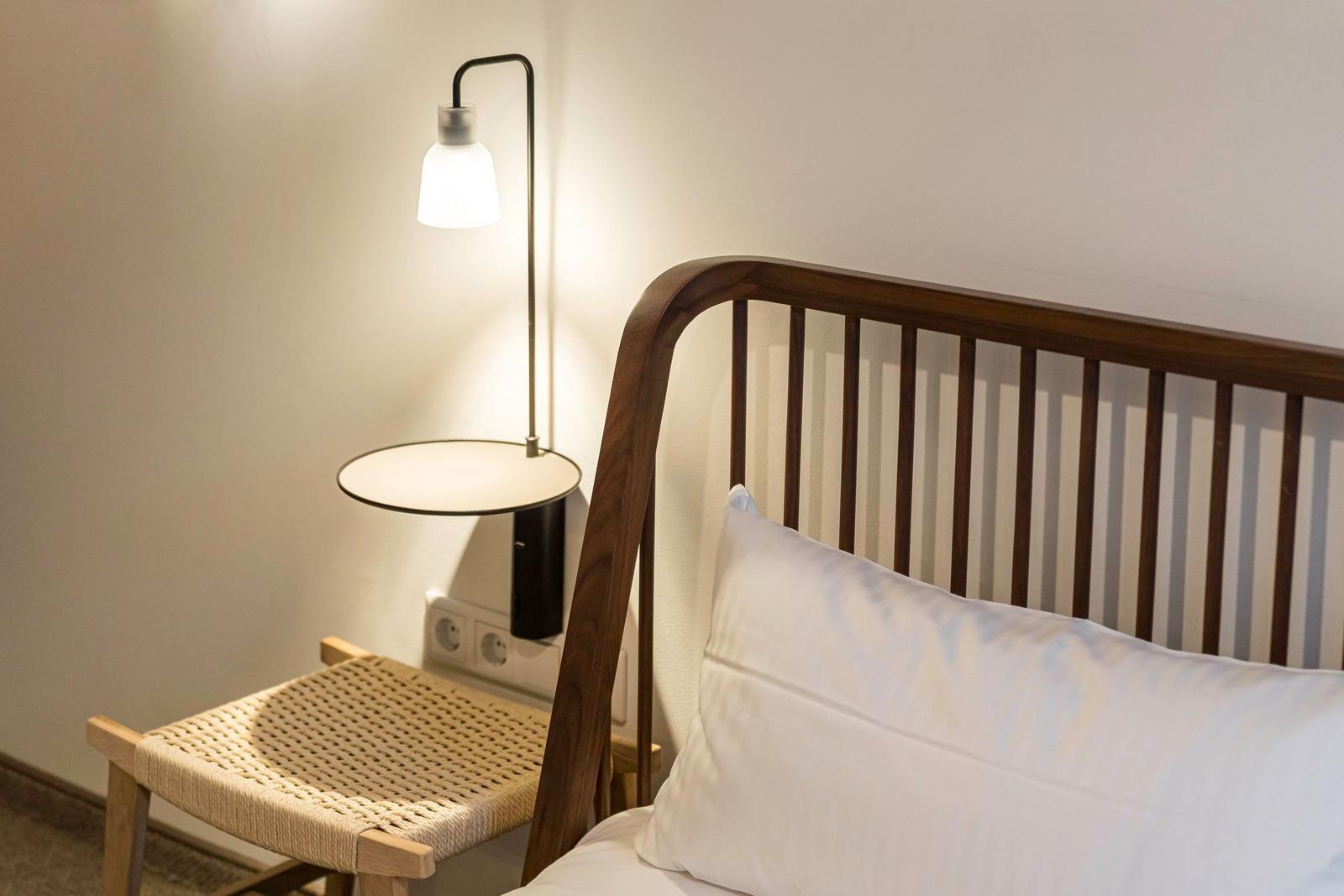 Ein Bett mit Holzgestell und weißer Bettwäsche neben einer Stehlampe und einem Stuhl.