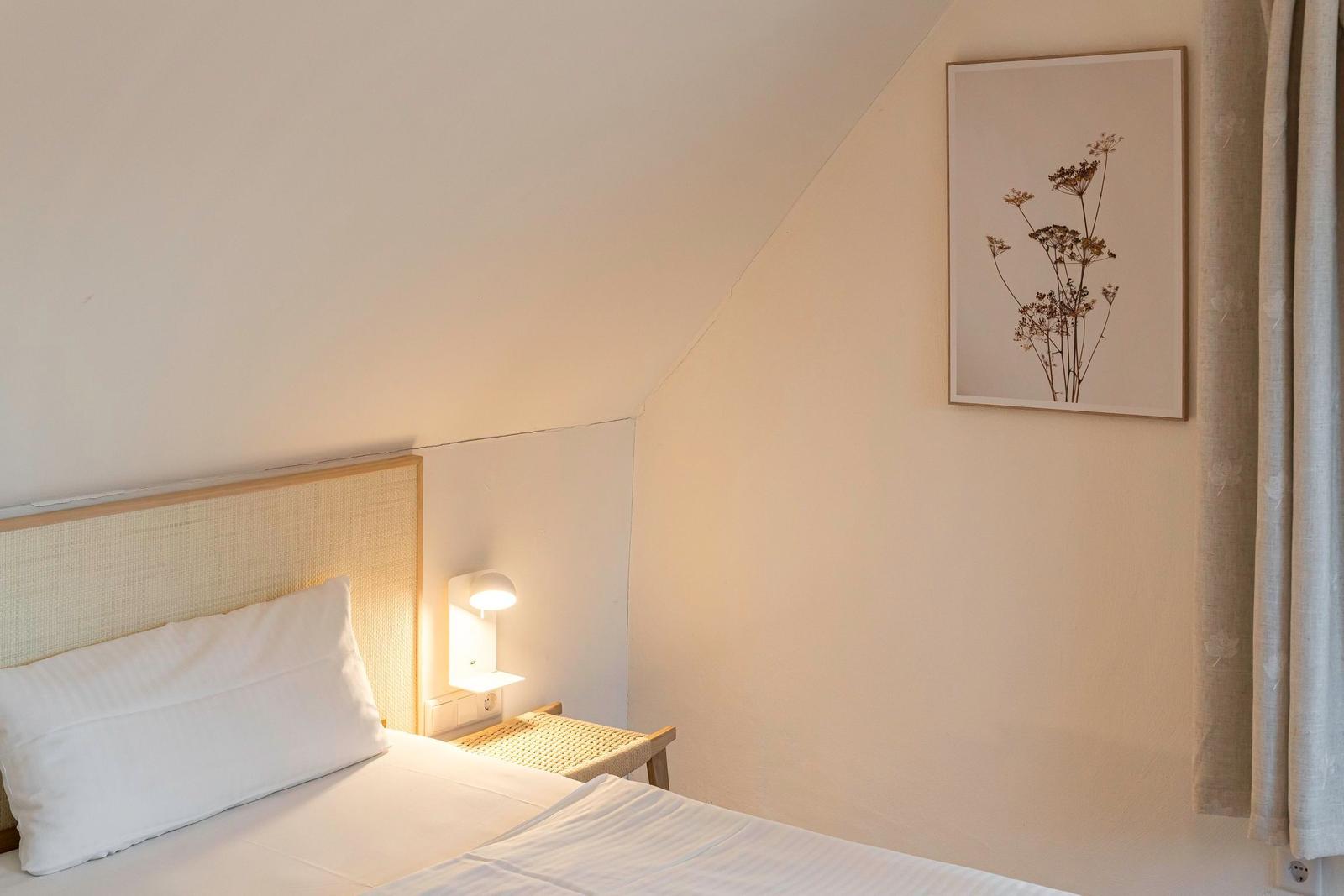 Schlafzimmer mit Bett, Nachttisch und Wandbild. Dachsatteldach und Vorhang sichtbar.