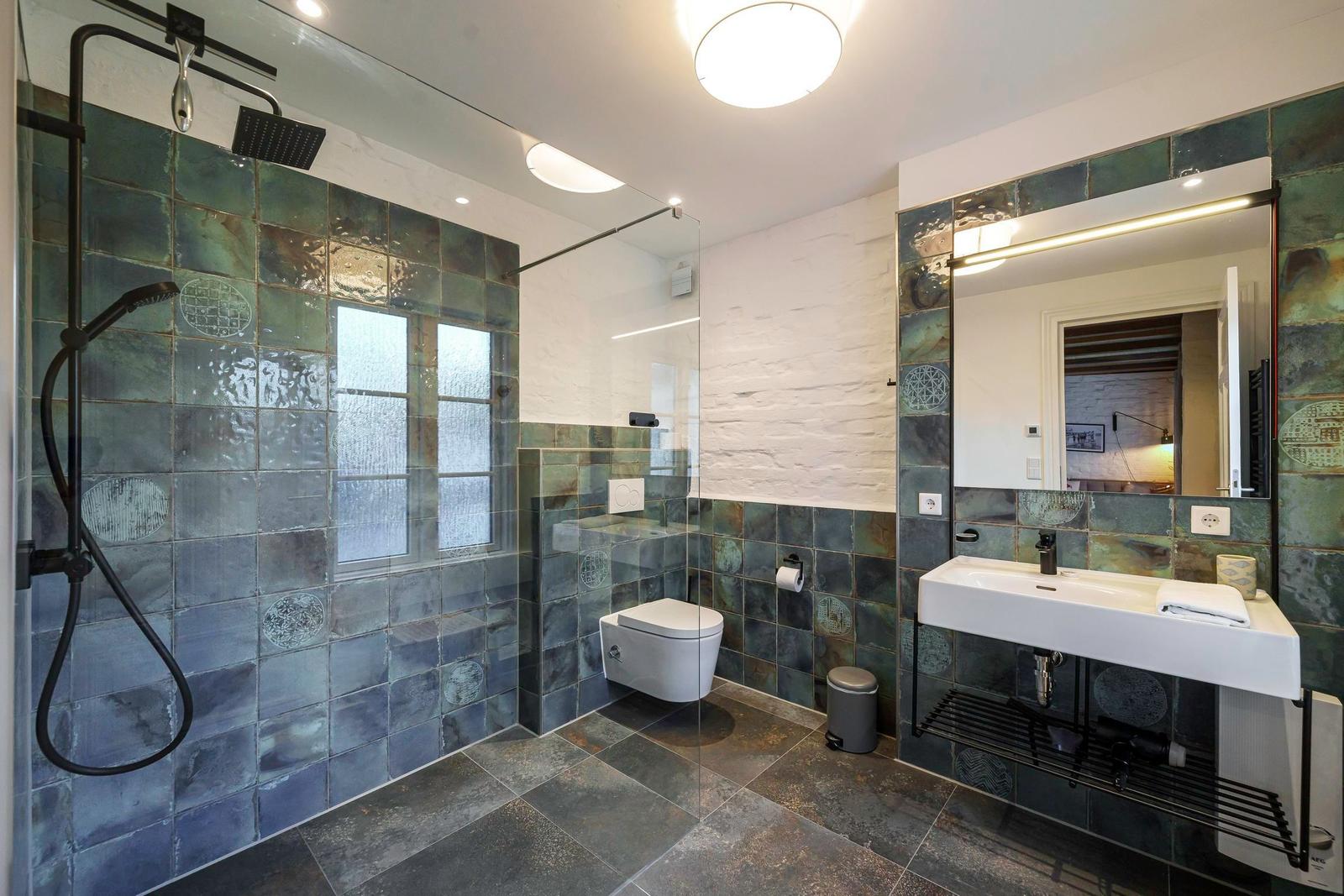 Bad mit Dusche, Waschbecken und Toilette. Wandfliesen in Blau- und Grüntönen.