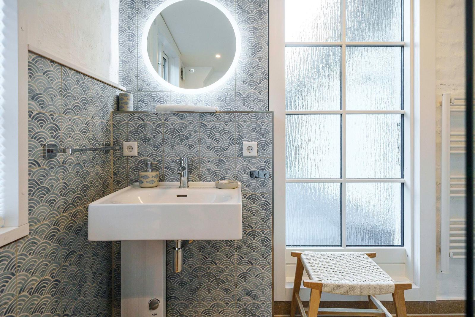 Bad mit Waschbecken, runden Spiegel und gefliesten Wänden. Fenster mit Sichtschutz und Holzstuhl.