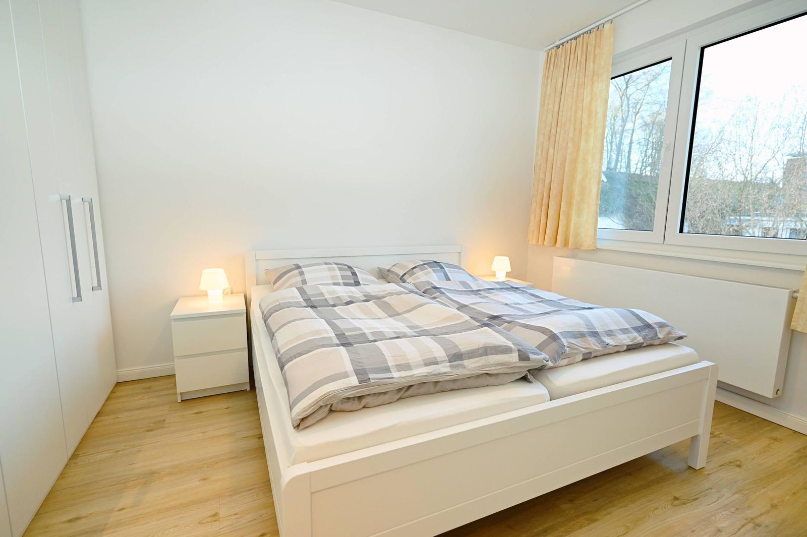 Schlafzimmer mit Doppelbett, Nachttischen und Fenster. Holzfußboden und weiße Möbel.