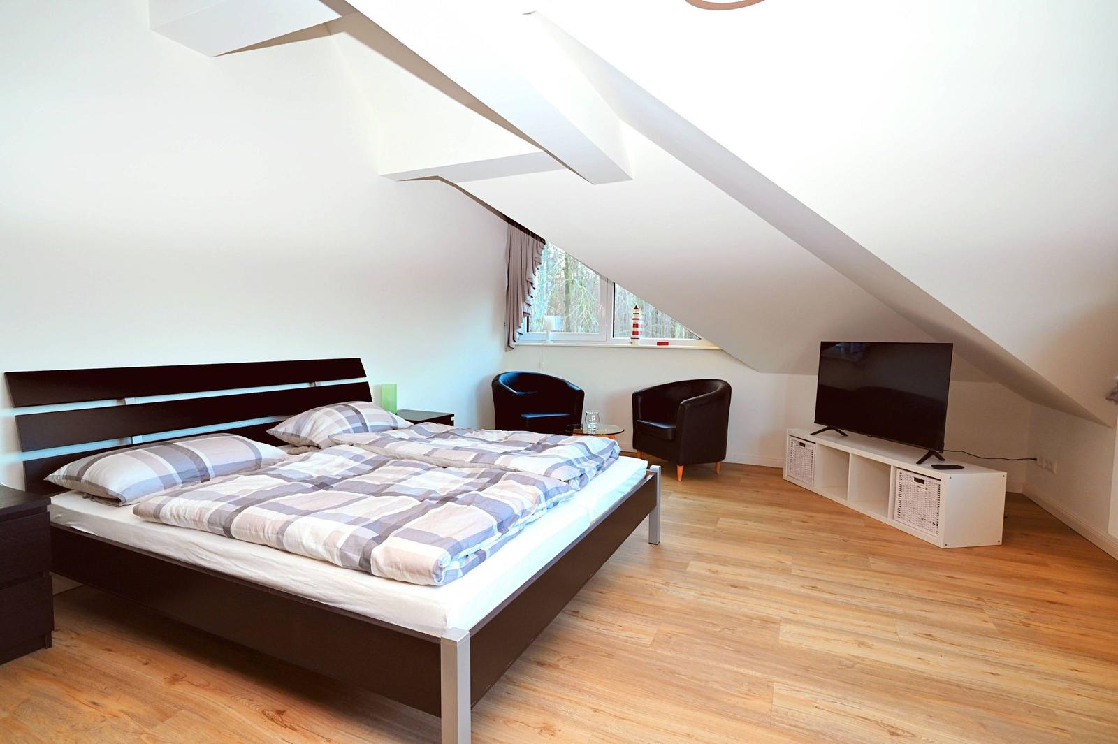 Schlafzimmer mit Dachschrägen, Bett, TV und zwei Sesseln.