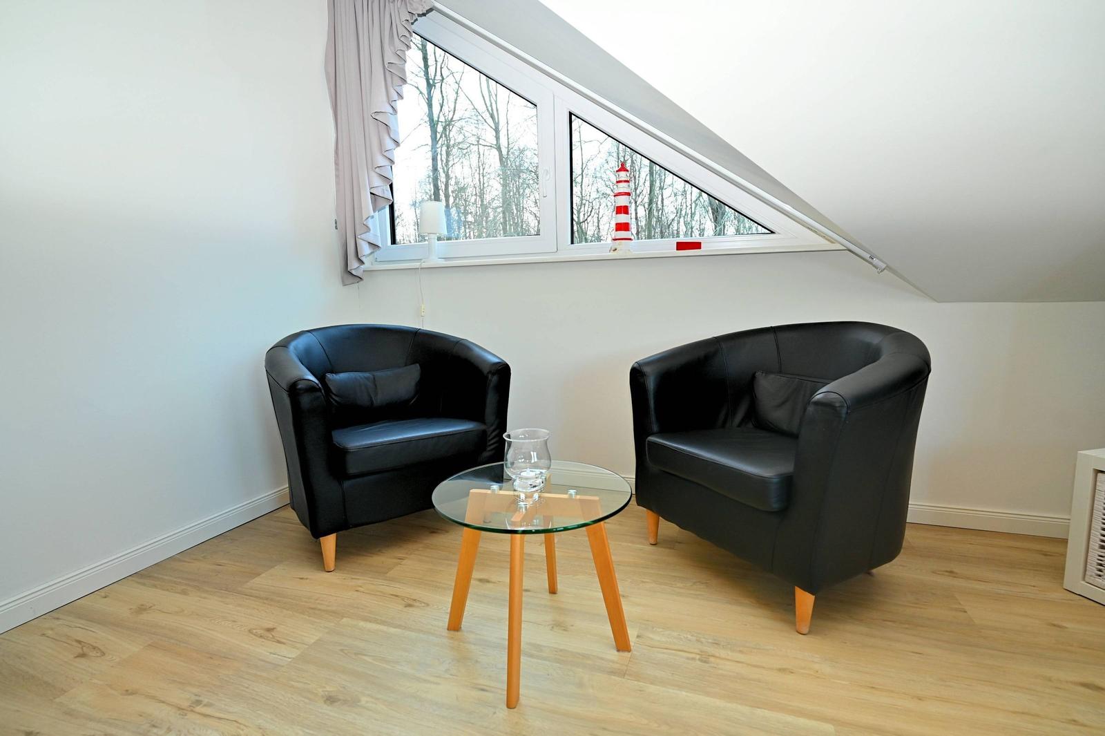 Zwei schwarze Sessel und ein Glas-Tisch stehen vor einem Dachfenster.
