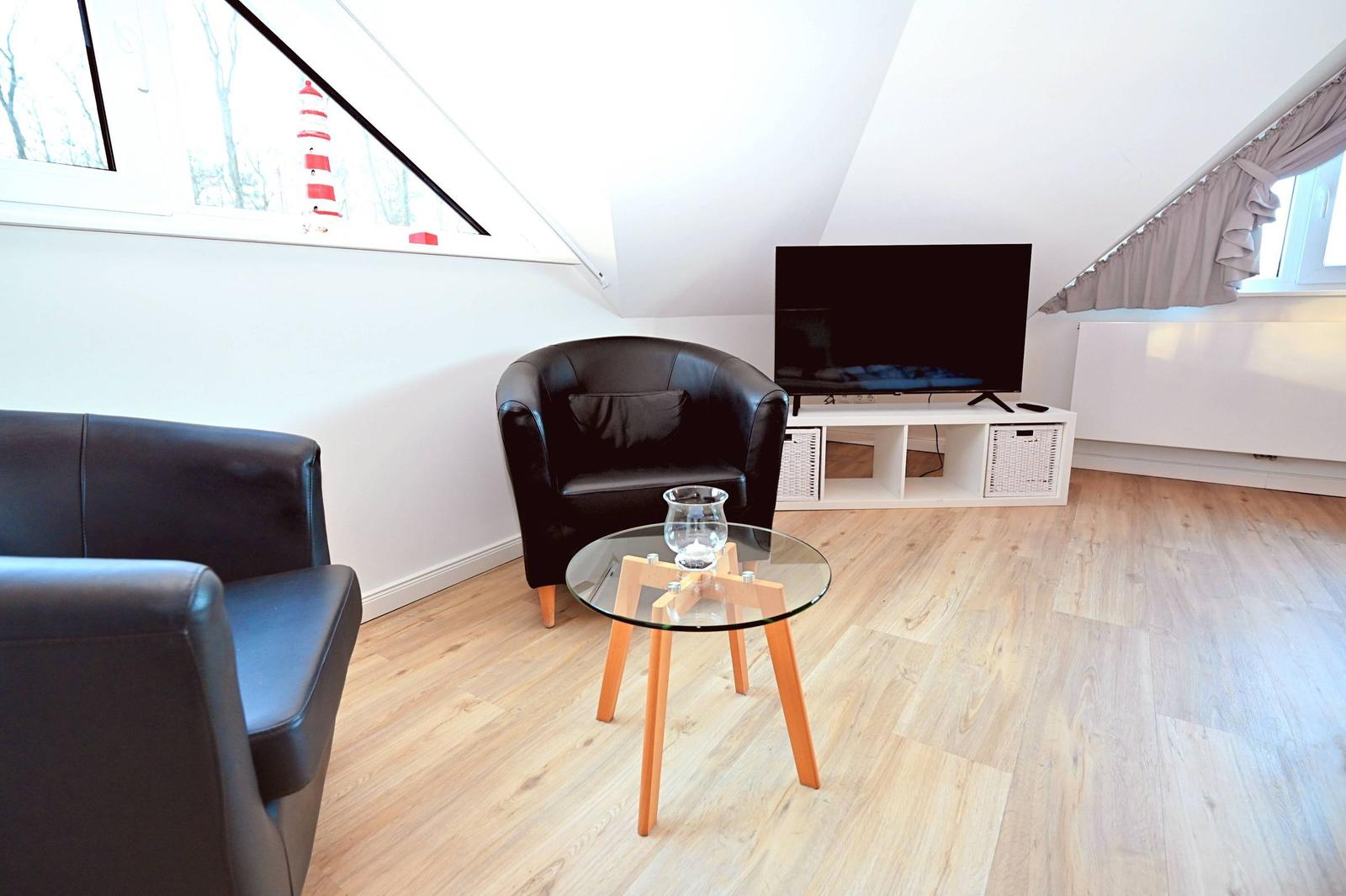 Wohnzimmer mit Sofa, Sessel, Glas-Tisch und Fernseher unter Dachschrägen.