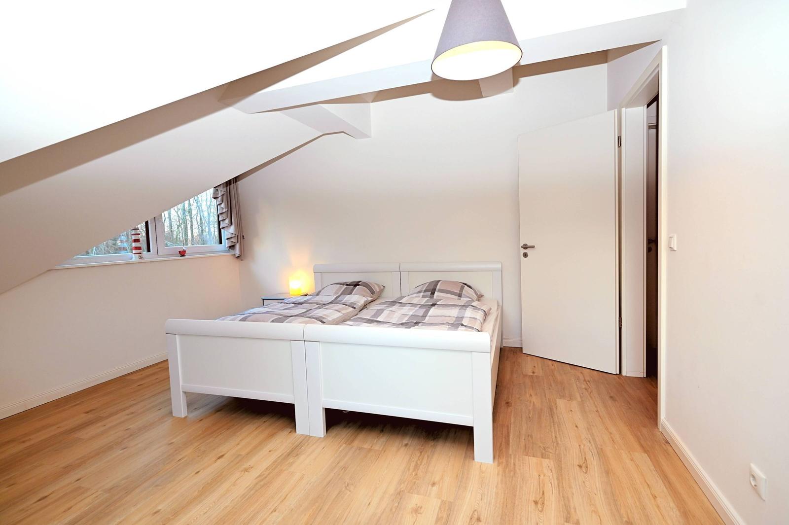 Schlafzimmer mit Dachschrägen, weißem Bett und Holzboden. Fenster mit Blick nach draußen.