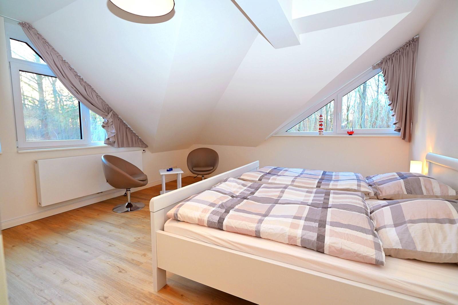 Schlafzimmer mit Dachschrägen, Bett, zwei Sessel und Fenstern mit Blick auf Wald.