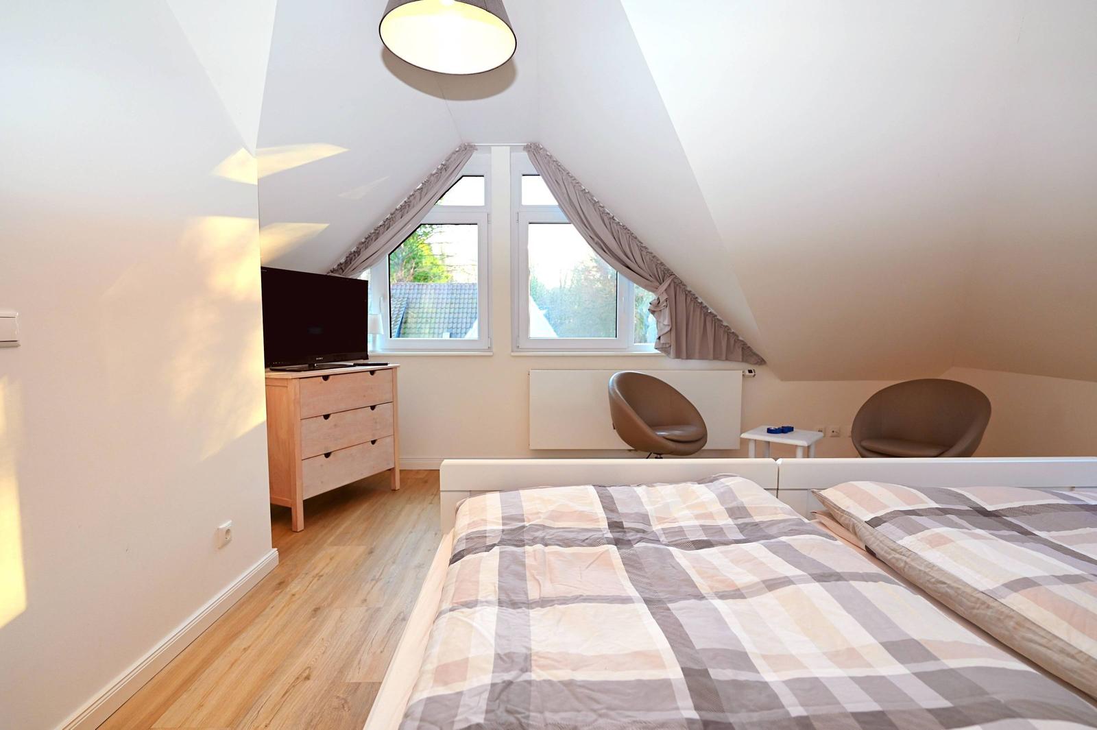 Schlafzimmer mit Doppelbett, zwei Sesseln, TV und Fenster mit Blick auf Dächer.