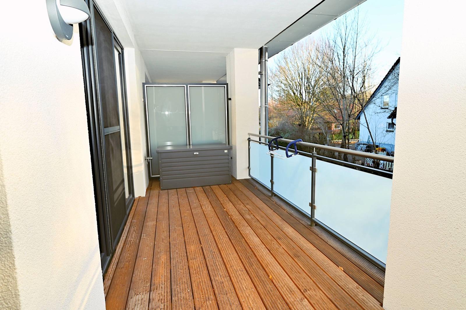Terrasse mit Holzdielen, Glasgeländer und Außenspeicher. Blick auf Bäume und Nachbarhäuser.