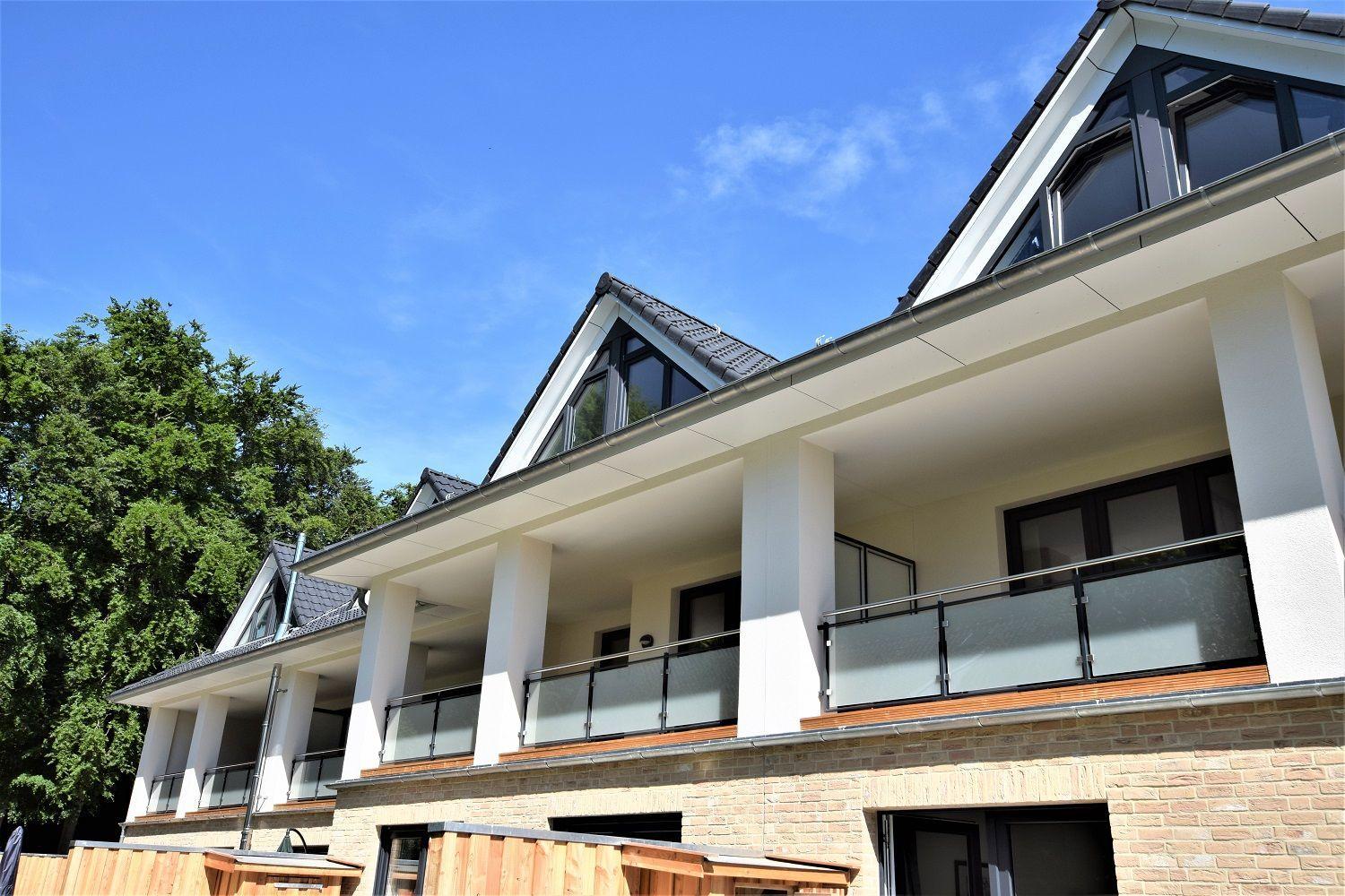 Moderne Villa mit Balkon und Dachfenstern, umgeben von Bäumen unter blauem Himmel.