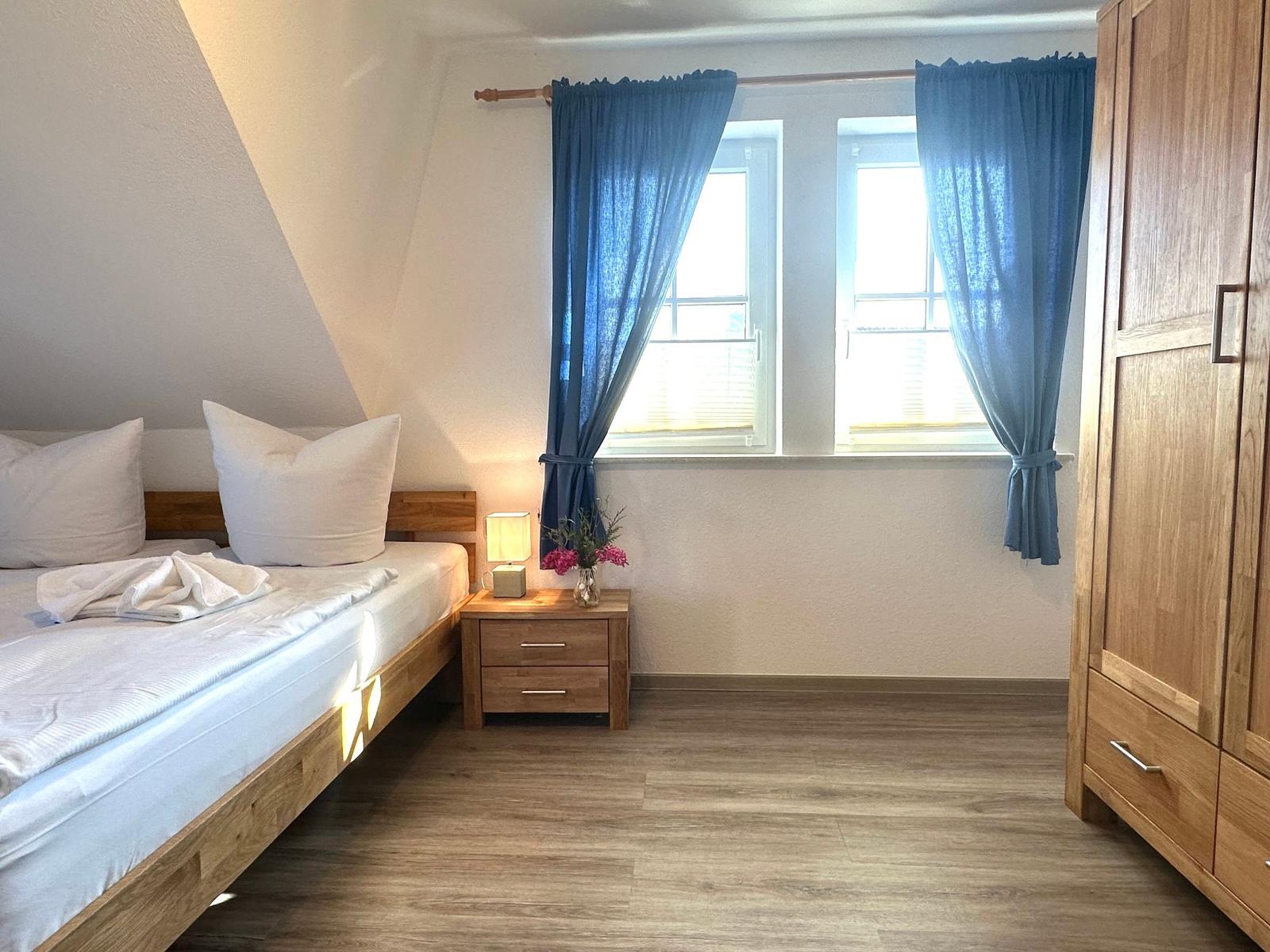Schlafzimmer mit Holzbett, Nachttisch und Kleiderschrank. Fenster mit blauen Vorhängen.