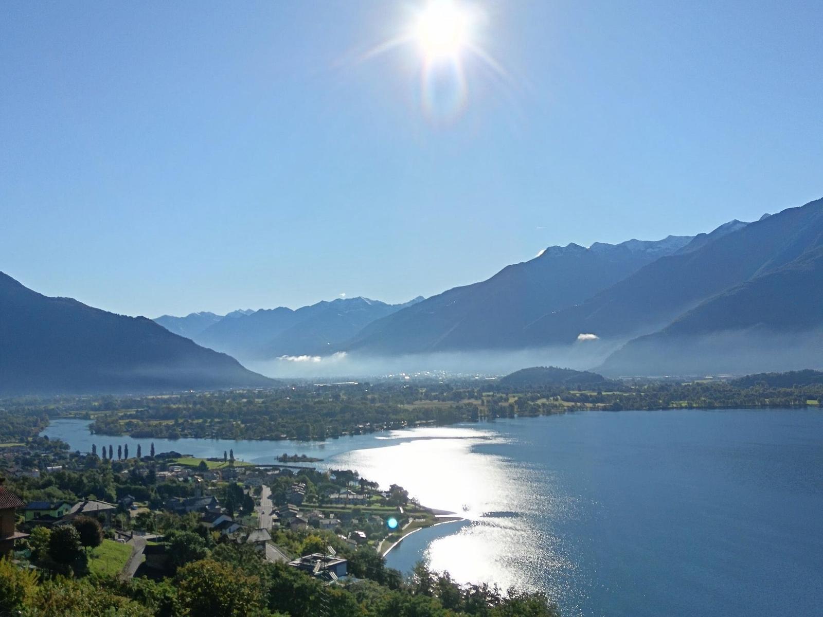 Panoramablick auf einen See mit Bergen im Hintergrund und Sonne am Himmel.