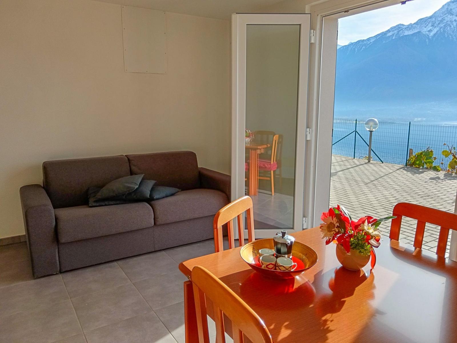 Wohn- und Essbereich mit Blick auf See und Berge. Holztisch, Sofa und Terrasse.