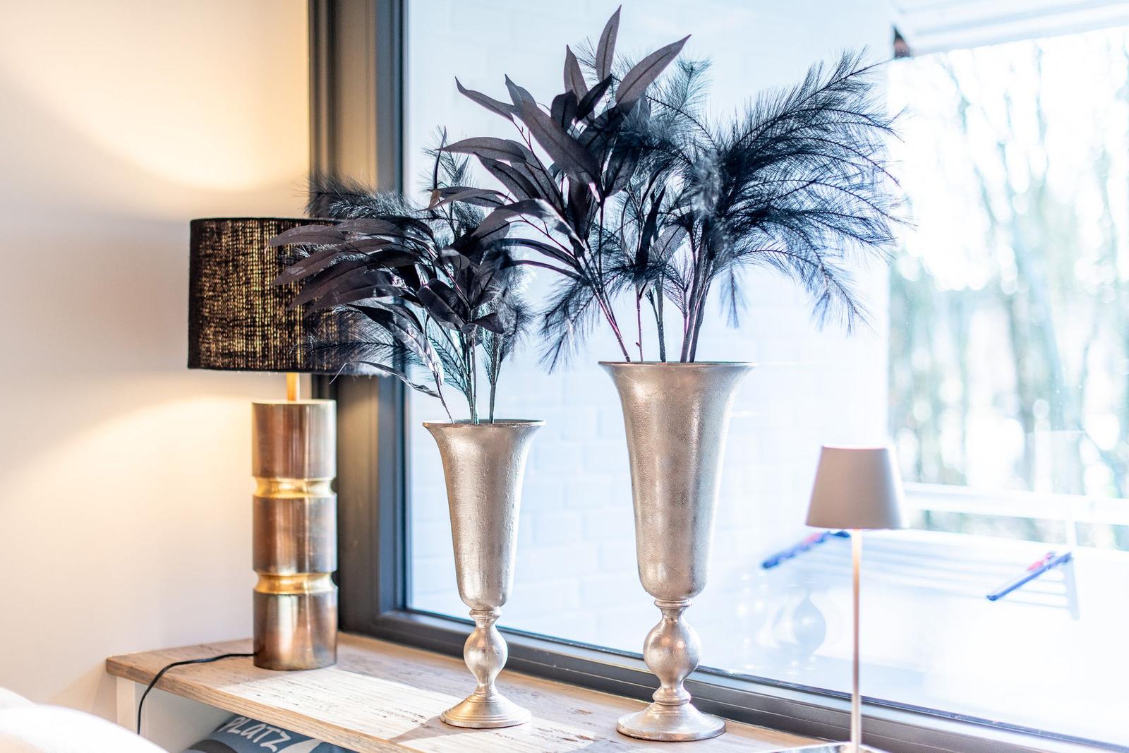 Zwei silberne Vasen mit dunklen Blättern stehen auf einem Holzregal neben einem Fenster.