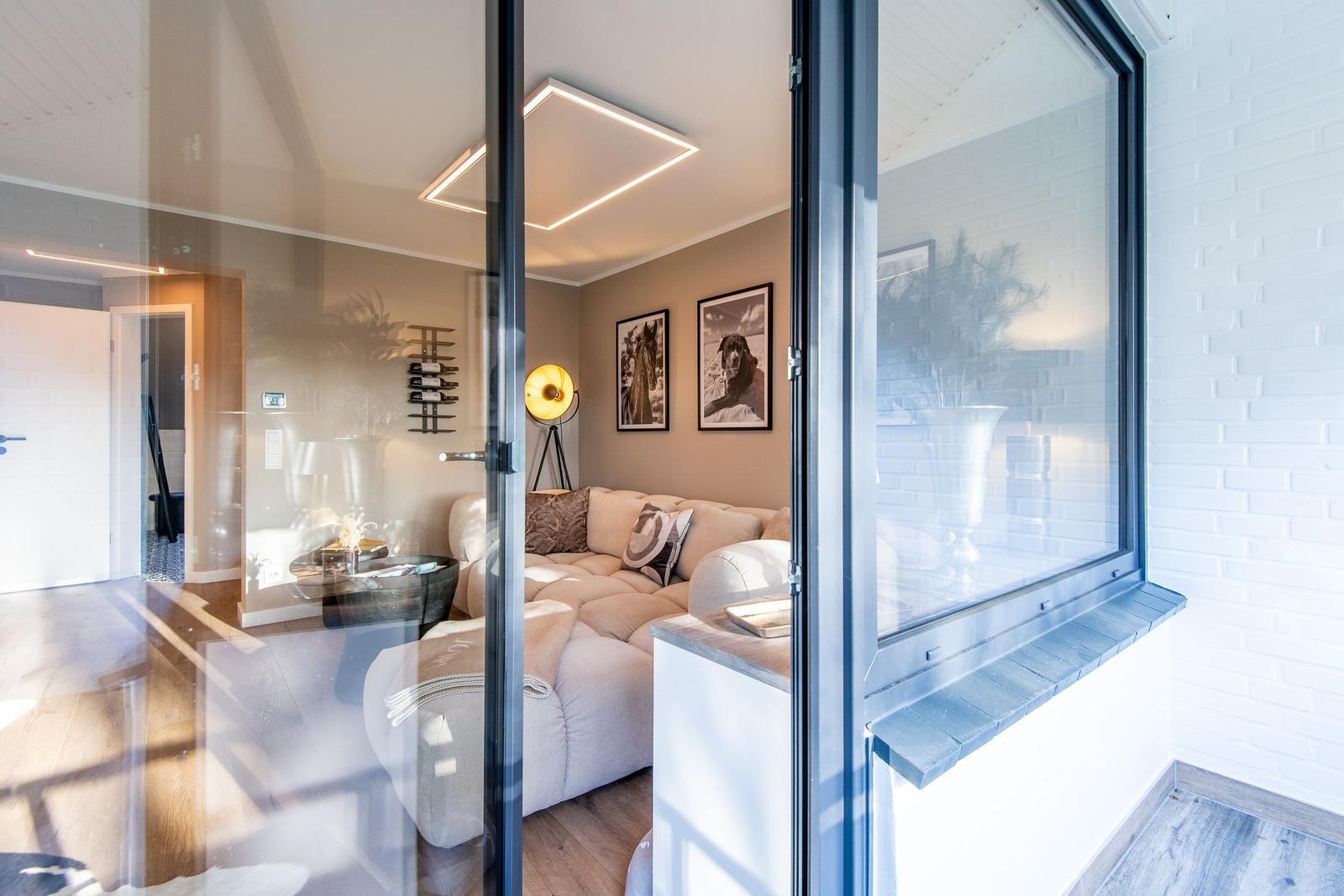 Durchgehendes Glas führt in ein Wohnzimmer mit Sofa, Wandbildern und einer Stehlampe.