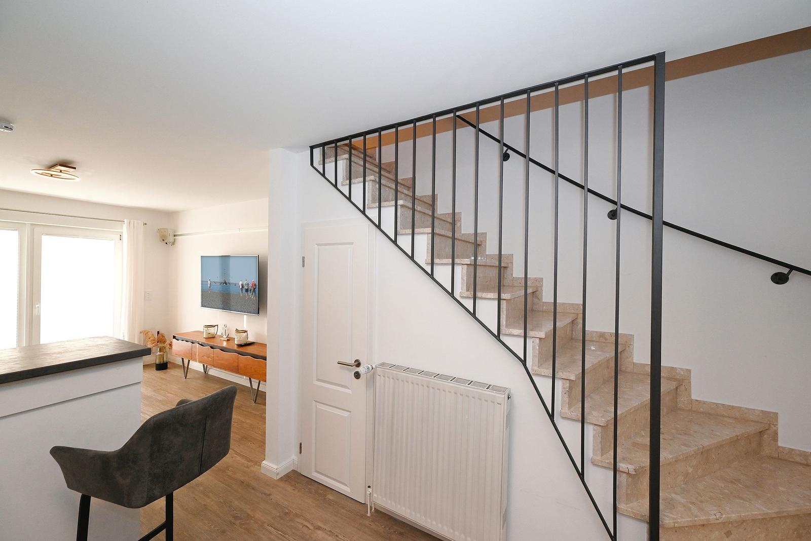 Treppe mit Metallgeländer führt zur oberen Etage. Im Hintergrund Wohnbereich mit Fernseher und Holzboden.