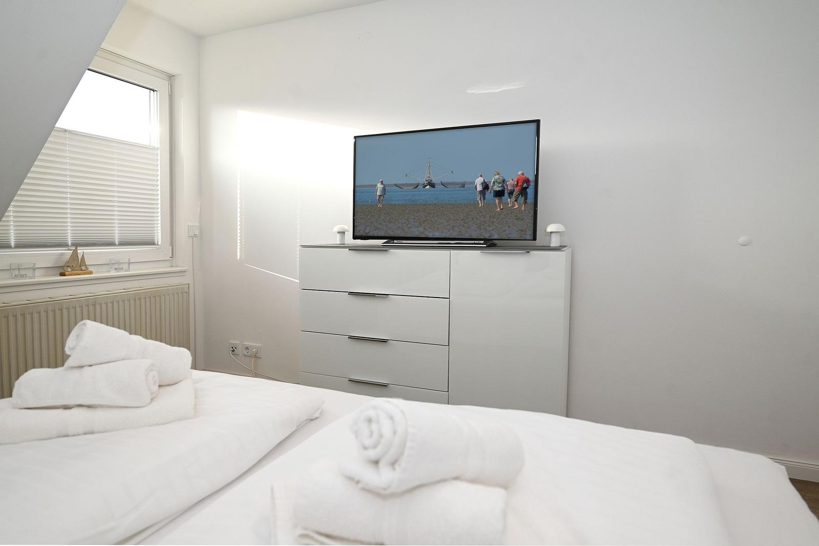 Schlafzimmer mit weißem Bett, Fernseher und Fenster mit Jalousie.